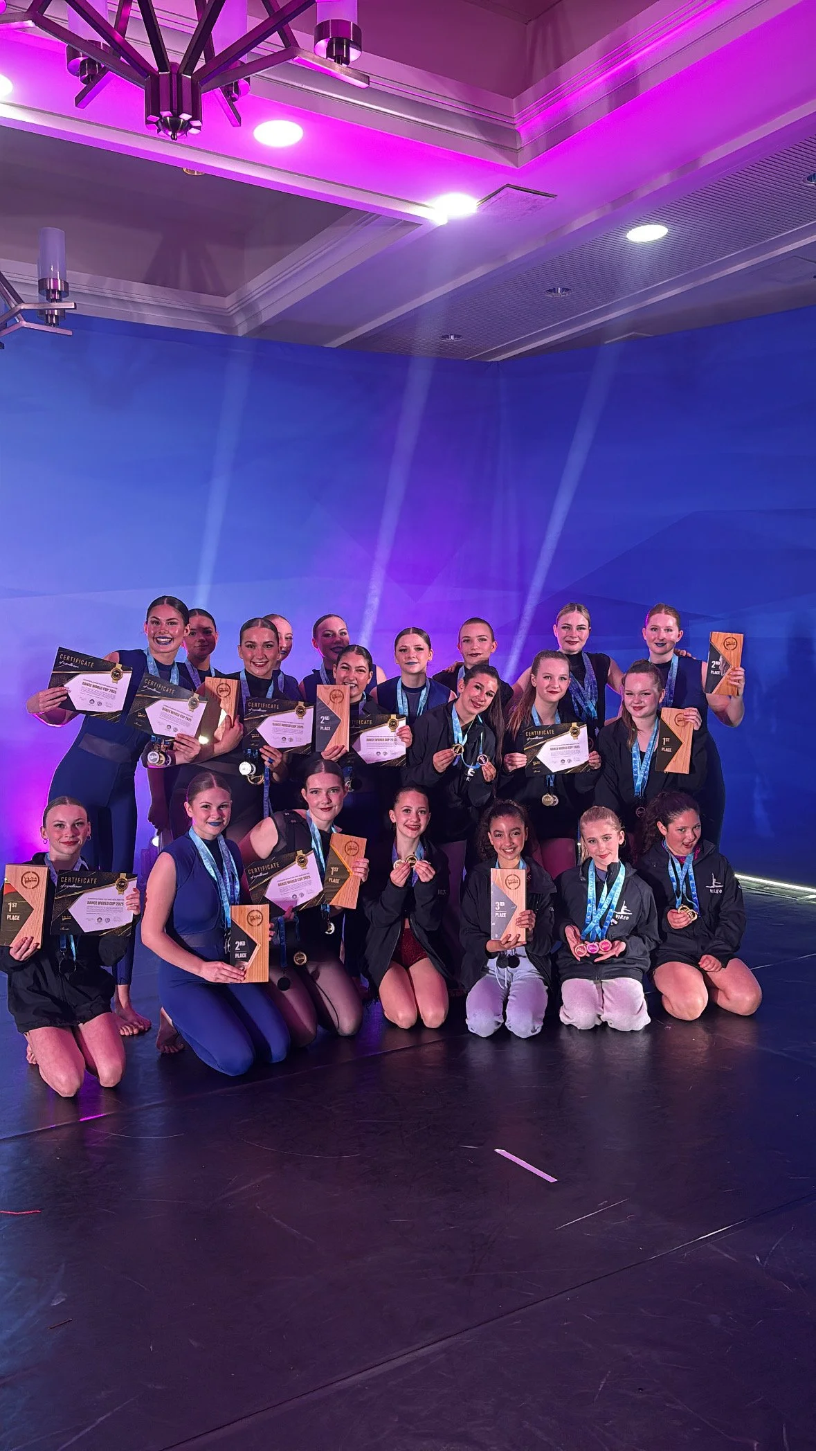 Group of young gymnasts on stage holding certificates, medals, and trophies at an awards ceremony