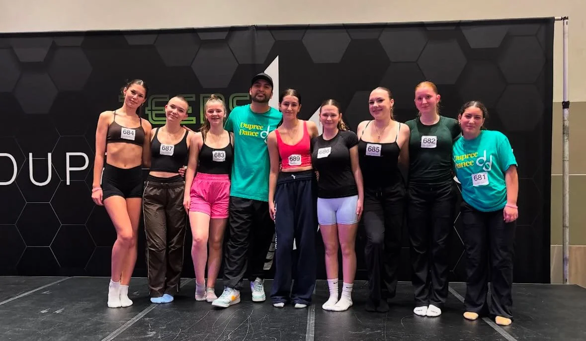 Group of eight dancers posing on stage in front of a black backdrop with hexagon patterns at a dance event