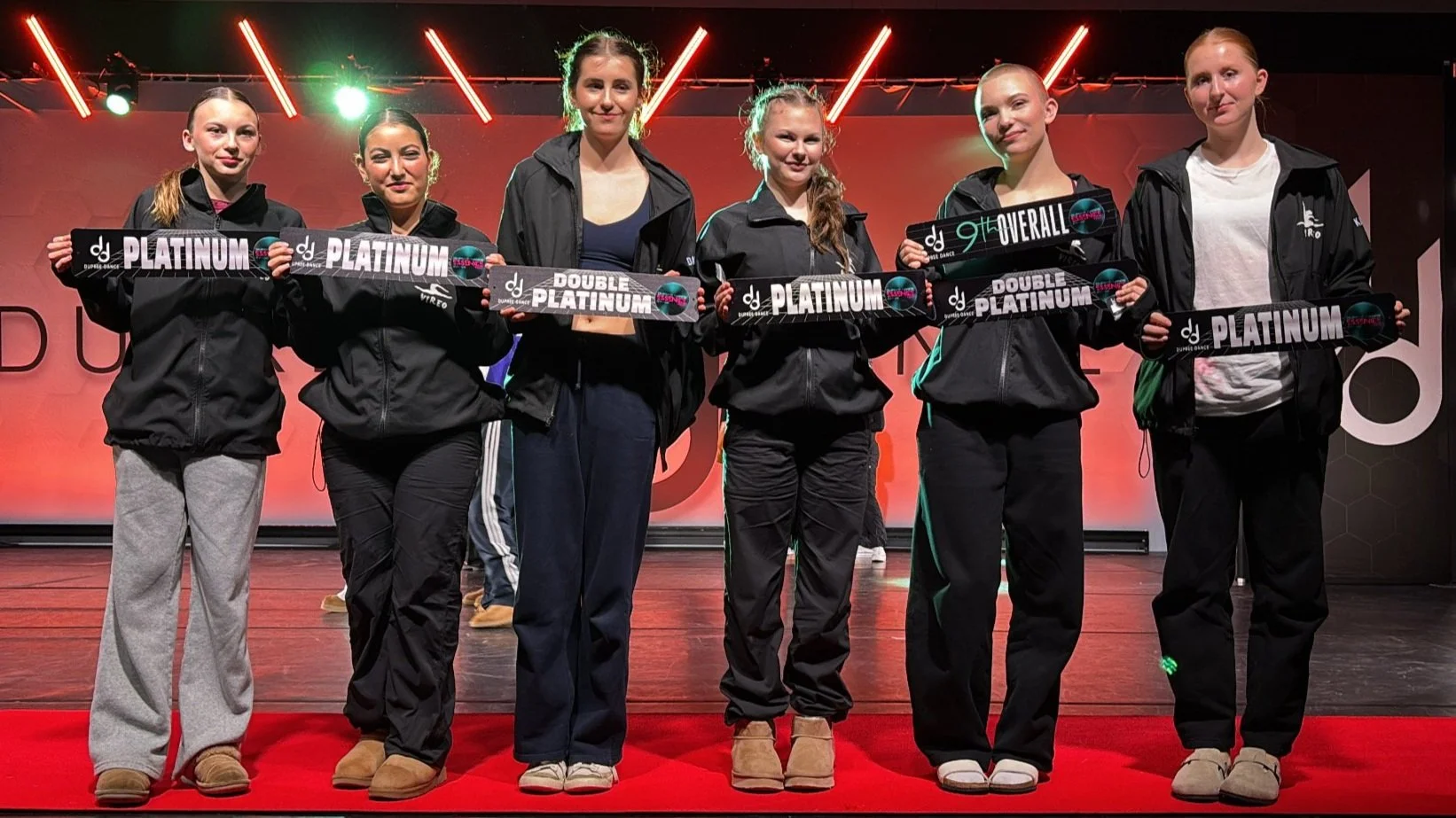 Six young women in black jackets holding awards including Platinum and Double Platinum on a stage with red lighting