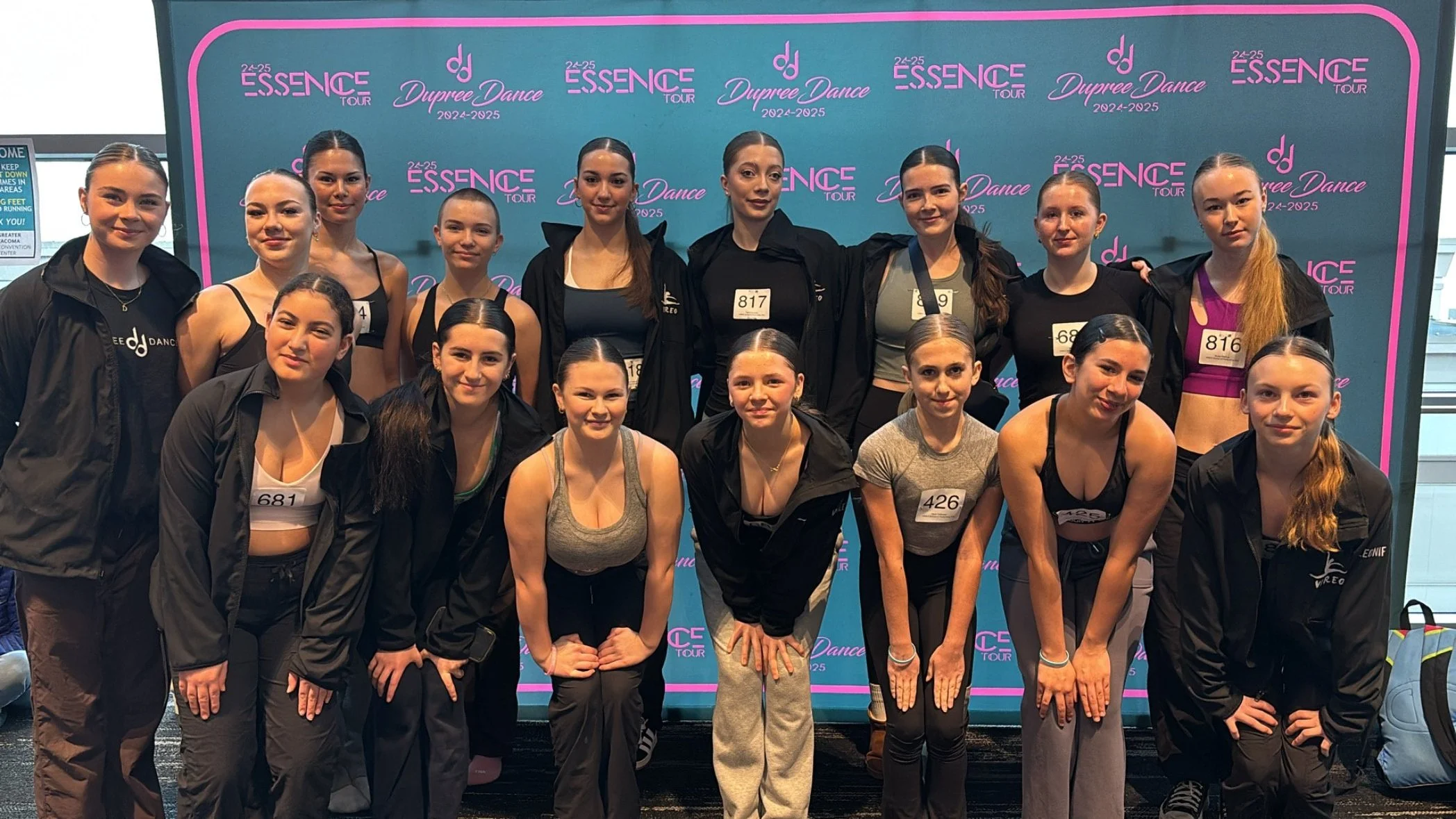 Group of young female dancers in dancewear and jackets posing in front of an Essence Tour backdrop at a dance competition