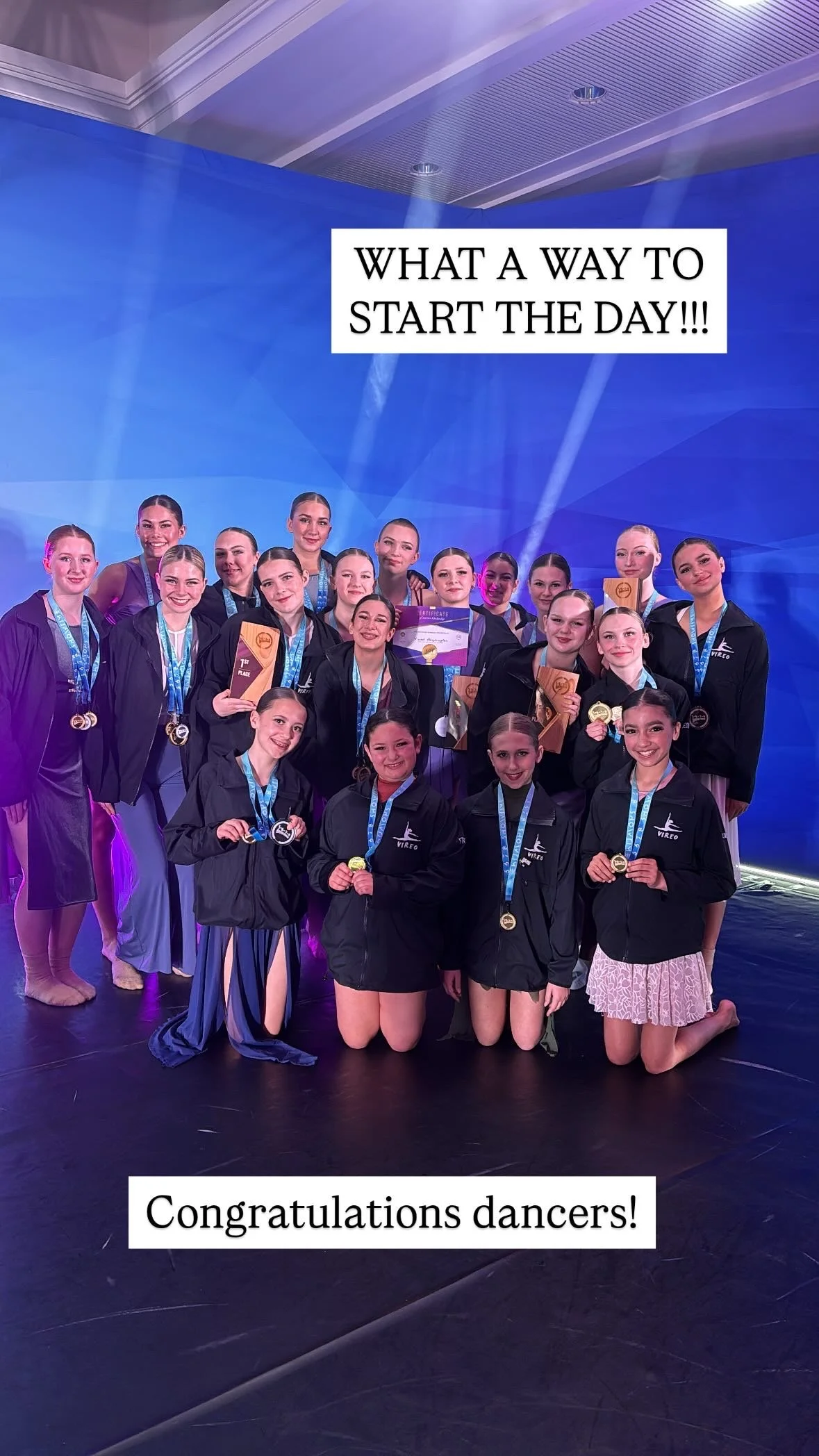 Group of young dancers with medals and holding awards smiling at the camera with blue backdrop and stage lights