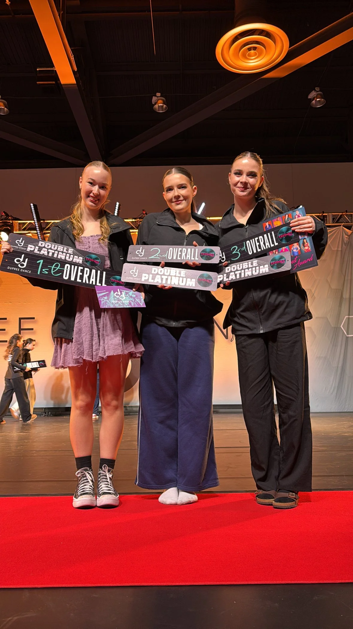 Three young women standing on a stage holding awards for dance competition overall placement first second and third