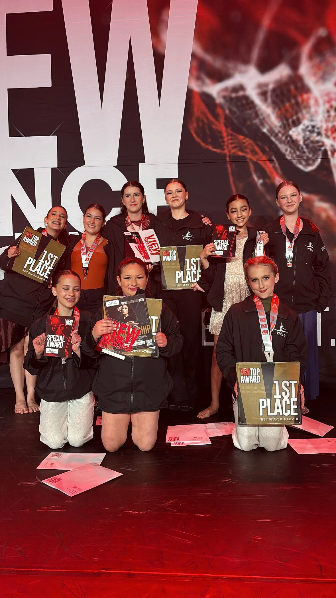 A group of young female dancers on stage holding awards and certificates with large screen displaying VIEW